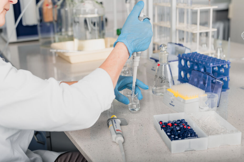 Na zdjęciu widać prace w laboratorium. Dłonie ubrane w rękawiczki lateksowe obsługują automatyczna pipetę.