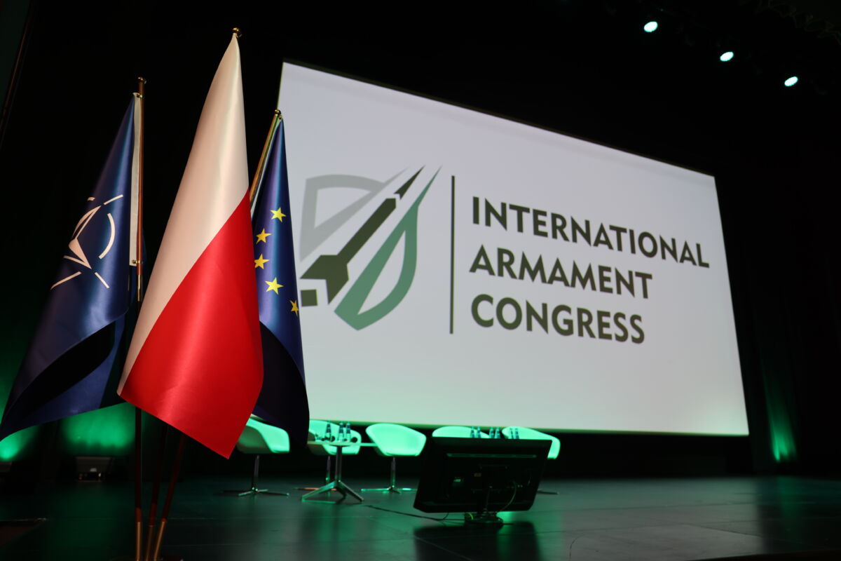 Scena podświetlona na zielono. Na pierwszym planie widnieją flagi Unii Europejskiej, Polski oraz NATO. W tle widać wyświetlony napis International Armament congress.